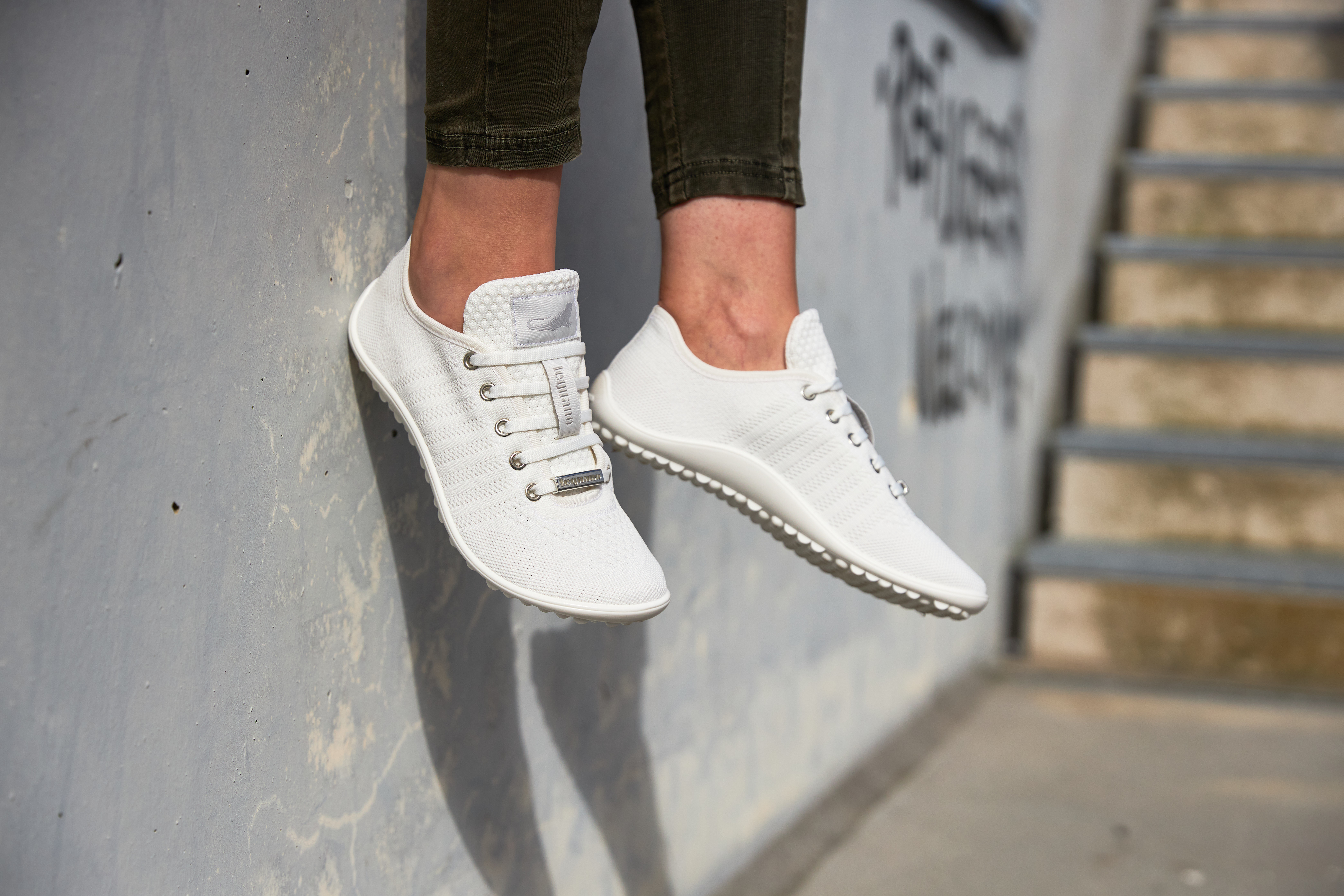 Zwei Füße in weißen Barfußschuhen vor einer Betonwand, rechts daneben eine Treppe