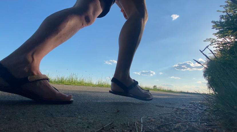 Beine und Füße eines Läufers in Barfußsandalen auf einem Feldweg vor blauem Himmel